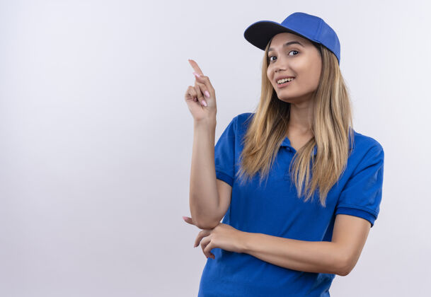 微笑面带微笑的年轻送货员身穿蓝色制服 戴着帽子 指着隔离在白色墙上的一面 留着复印空间穿女孩制服