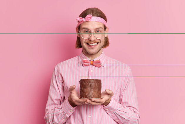 男性快乐的白种人去庆祝生日微笑着宽大地戴着头带衬衫和蝴蝶结拿着巧克力蛋糕和燃烧的蜡烛隔离在粉红色的墙上周年纪念庆祝室内
