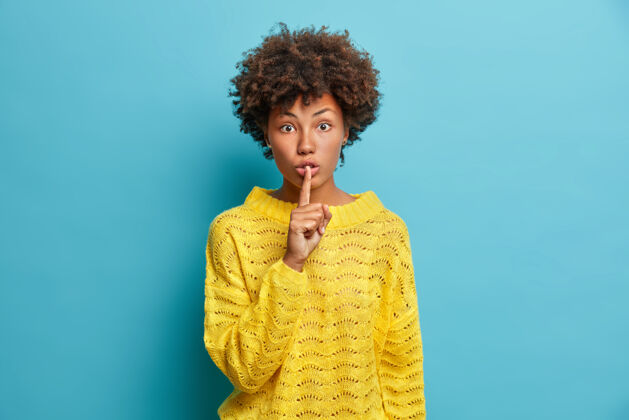 非洲惊讶的年轻女子做嘘手势要求保密显示嘘手势有震惊的表情穿着休闲的黄色毛衣隔着蓝色的墙八卦嘘室内