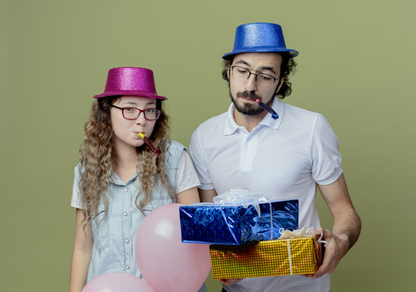橄榄一对戴着粉色和蓝色帽子的年轻夫妇吹着口哨 一个男人拿着礼物盒 看着隔离在橄榄绿墙上的礼物盒帽子小伙子吹口哨