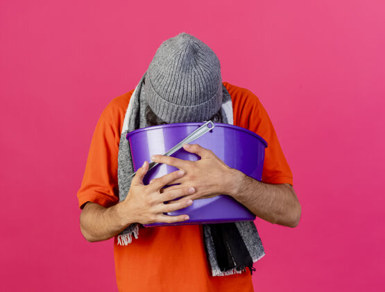 男人年轻的白人病人戴着眼镜 戴着冬天的帽子和围巾 手里拿着塑料桶 恶心呕吐 把桶隔离在深红色的墙上呕吐塑料疾病