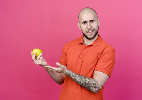 不愉快一个不开朗的年轻人手拿苹果 指着隔离在粉红色墙上的苹果手拿着年轻