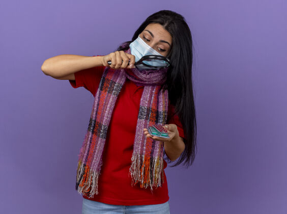 胶囊戴着口罩和围巾的年轻白种人女孩拿着一包包胶囊 通过放大镜看着它们 隔离在紫色的墙上 留着复印空间疾病穿过年轻