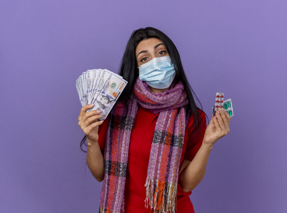 钱自信的年轻白种女孩戴着面具 戴着围巾 手里拿着钱和几包胶囊 隔离在紫色的墙上 留着复印空间围巾胶囊年轻