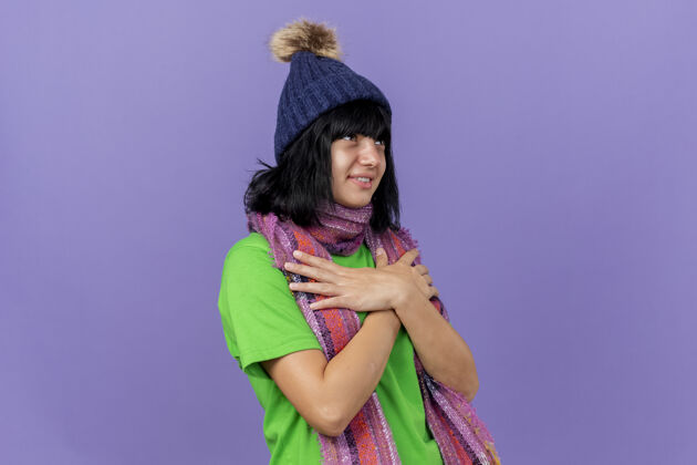 年轻的戴着冬季帽子和围巾的年轻白种女孩高兴地抬起头来 双手交叉放在胸前 隔离在紫色的墙上 留着复制空间保存十字架围巾