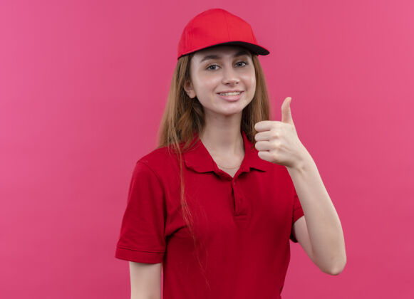 女孩微笑着的年轻送货女孩 穿着红色制服 在粉红色的墙壁上竖起大拇指 留着复印空间年轻微笑女人