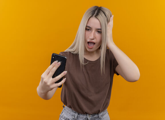 年轻惊讶的年轻金发女孩拿着手机看着它 手放在头上孤立的橙色墙壁与复制空间抱着女人金发
