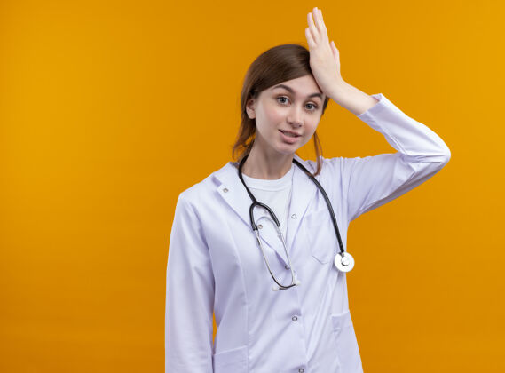 女人惊讶的年轻女医生穿着医用长袍和听诊器把手放在头上孤立的橙色墙壁与复制空间手女人医生