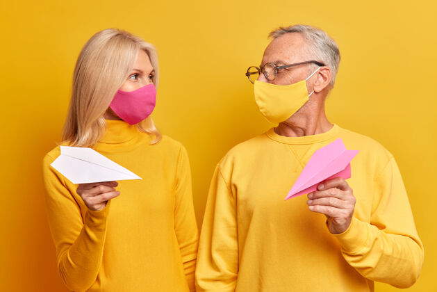 眼镜成熟的女人和男人开心地看着对方戴着防护面具摆姿势用手工制作的纸飞机尽量保持距离 防止冠状病毒在黄色墙壁上隔离传播成熟流行病毒