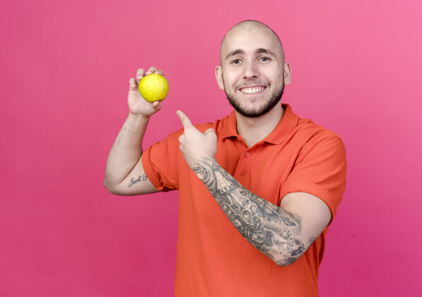 男人面带微笑的年轻运动型男子拿着苹果指着粉红色墙上的孤立点苹果微笑
