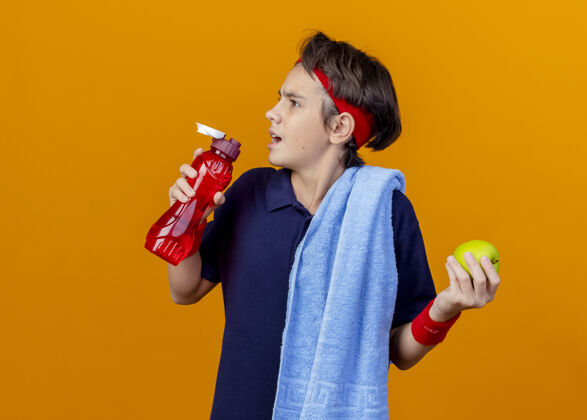 壁板年轻帅气的运动男孩戴着头带和护腕 肩上戴着牙套和毛巾 头转向一边 拿着苹果和水瓶 隔离在橙色的墙上 留着复制空间头带橙色牙齿