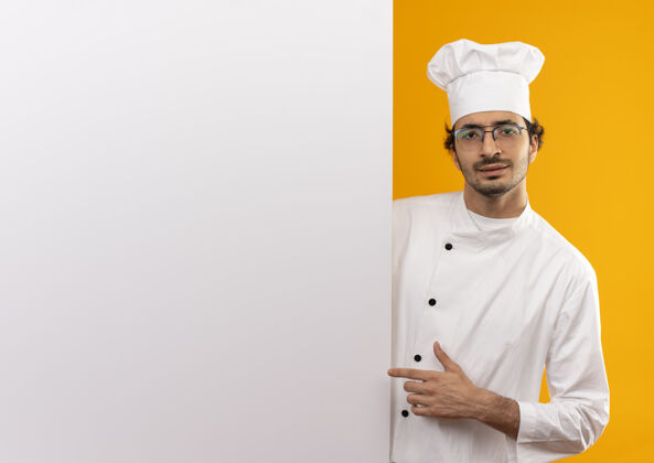 不愉快年轻的男厨师穿着厨师制服 戴着眼镜 拿着白色的墙 隔离在黄色的墙上 留着复印空间眼镜穿着墙