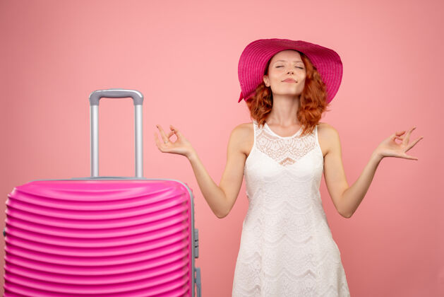 美丽年轻女性游客戴着粉色帽子和包在粉色墙壁上冥想的正面图前面人包