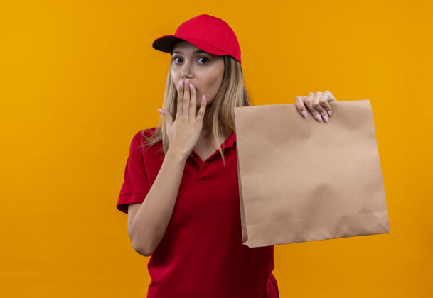 拿着被吓坏了的年轻送货女孩穿着红色制服 戴着帽子 手里拿着纸袋 嘴巴被隔离在橙色的墙上送货女孩年轻