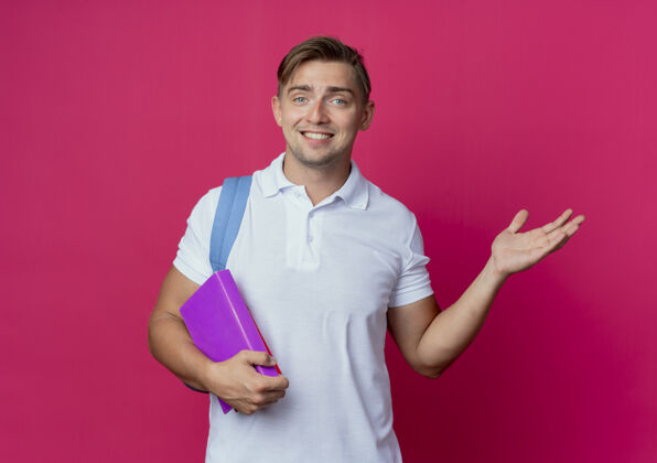 笑笑容可掬的年轻帅气男生背着书包拿着书点着手边隔离在粉红色的墙上年轻手男