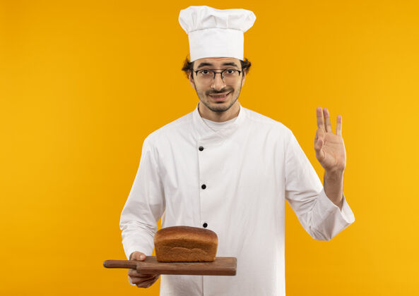 厨师年轻的男厨师穿着厨师制服 戴着眼镜 拿着面包放在砧板上 在黄色的墙壁上显示出很好的姿势眼镜板请