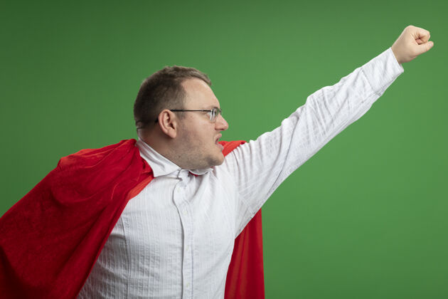 拳头自信的成年斯拉夫超级英雄 穿着红色斗篷 戴着眼镜 看着一边举起拳头隔离在绿色的墙上斯拉夫信心眼镜