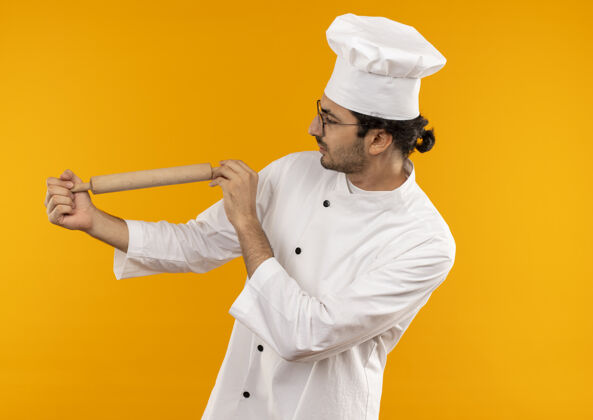 眼镜想着年轻的男厨师穿着厨师制服戴着眼镜拿着擀面杖看着隔离在黄色墙上的擀面杖卷男性年轻