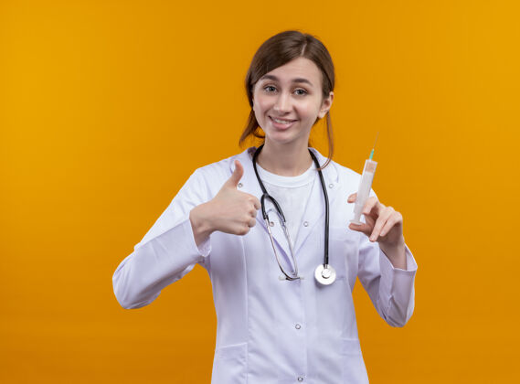 医生面带微笑的年轻女医生 穿着医用长袍 手持注射器 在隔离的橙色墙上竖起大拇指注射器年轻人女人