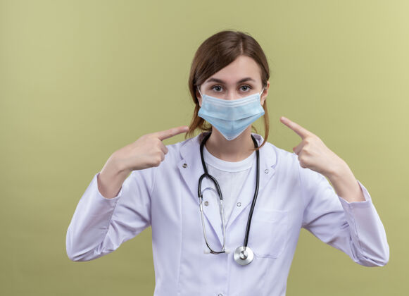 指尖年轻的女医生穿着医用长袍 戴着口罩 用听诊器指着隔离的绿墙上的口罩听诊器长袍口罩