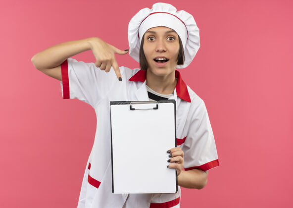 持有印象深刻的年轻女厨师在厨师制服举行 并指着剪贴板上的粉红色与复制空间隔离印象厨师烹饪