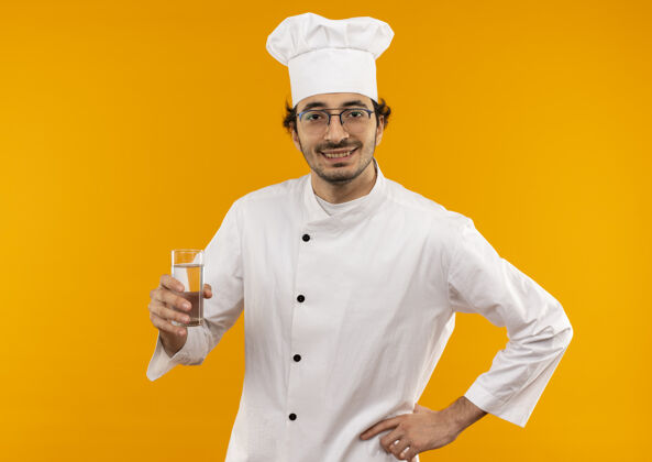 男性面带微笑的年轻男厨师穿着厨师制服 戴着玻璃杯 手放在黄色墙壁上的臀部烹饪手厨师