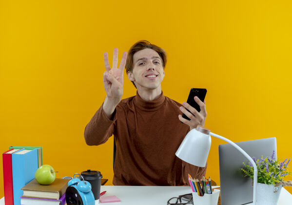 男孩微笑的年轻学生男孩坐在书桌旁 手里拿着学习工具 手里拿着电话 在黄色的墙上展示着三个孤立的孩子学生工具抱着