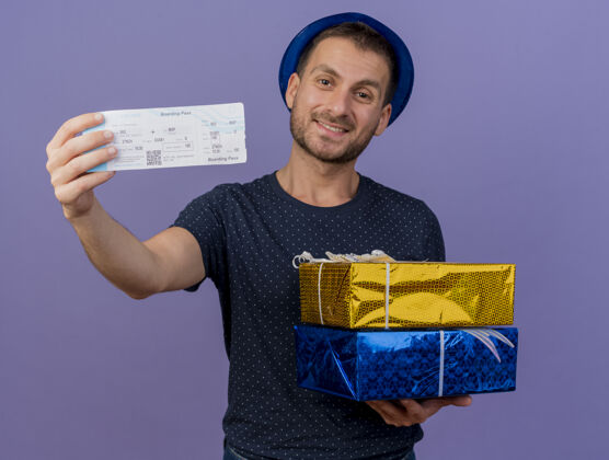 微笑面带微笑的白人帅哥戴着蓝色帽子拿着礼盒和机票 背景是紫色的 有复印空间持有航空礼物