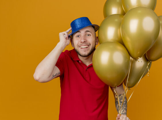 帅哥快乐的年轻英俊的斯拉夫党人戴着党的帽子拿着气球看着相机抓取橙色背景孤立的帽子帽子抓住抱着
