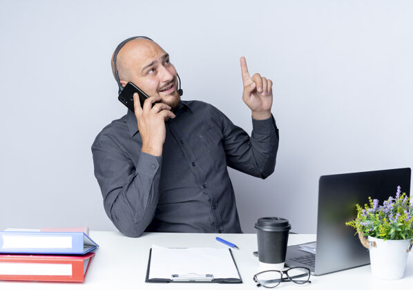 磨损印象深刻的年轻秃头呼叫中心男子戴着耳机坐在办公桌上与工作工具通话举起手指 看着隔离在白色的一边电话年轻印象
