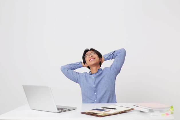 笔记本电脑戴着眼镜的快乐放松的亚洲年轻商人坐在工作场所 双手捂着头 闭着眼睛 对着白墙做梦成功工作微笑