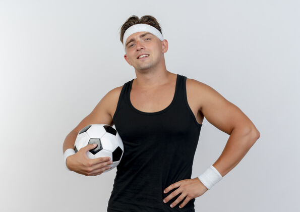 年轻自信的年轻帅气的运动型男人戴着头带和腕带 手里拿着足球 手放在腰上 隔离在白色衣服上腕带男人英俊