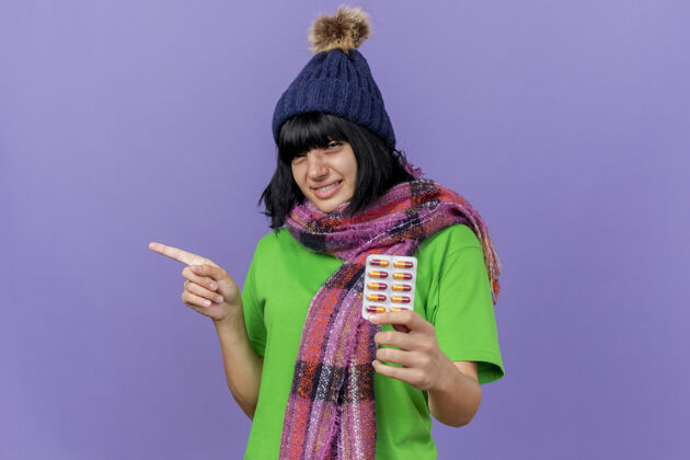 年轻人带着微笑的年轻病态的白人女孩戴着冬天的帽子和围巾 手里拿着一包胶囊 看着摄像机指着隔离在紫色背景上的一边 还有复印空间壁板微笑抱