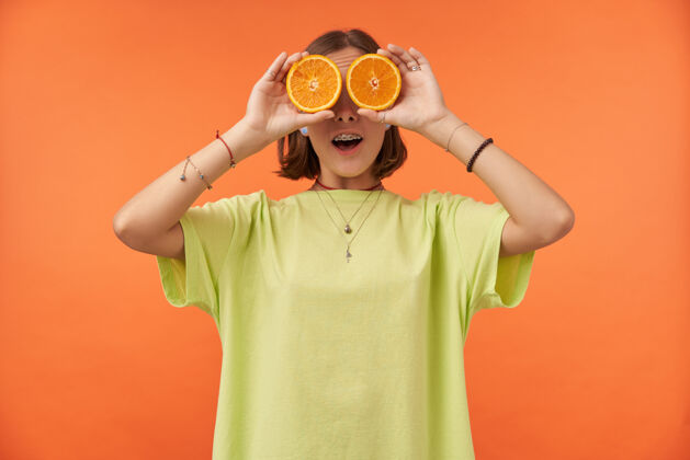 T恤女学生 一位棕发短发的年轻女士 眼睛上蒙着橘子神情惊讶站在橘色的墙上穿着绿色t恤 戴着牙套和手镯热带20多岁微笑