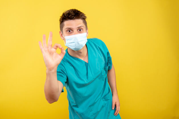男性正面图：年轻的男医生穿着医疗服 黄色背景上带着无菌口罩人医院医学
