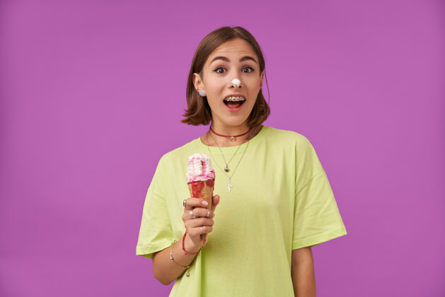 模特一幅迷人 震惊的女孩的画像 手里拿着冰淇淋 鼻子上放着冰淇淋穿着绿色t恤 戴牙套 戒指 项链和手镯女孩年轻人成人