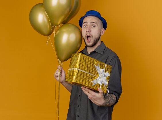 礼物惊讶的年轻英俊的斯拉夫党人戴着党的帽子站在侧视图气球和礼品包在橙色背景与复制空间看相机隔离年轻帽子举行