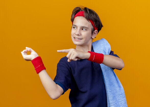 微笑面带微笑的年轻英俊的运动男孩戴着头带和护腕 肩上戴着牙套和毛巾 看着并指着橙色背景上孤立的一面运动壁板毛巾