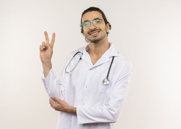 听诊器微笑的年轻男医生 戴着眼镜 穿着白色长袍 带听诊器 在隔离的白色墙壁上展示着和平的姿态 还有复印空间年轻穿和平
