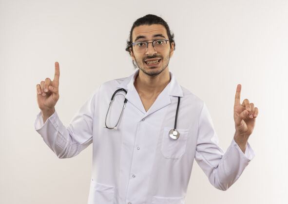 光学面带微笑的年轻男医生戴着眼镜 身穿白色长袍 手持听诊器 指着隔离的白色墙壁上有复印空间听诊器男性眼镜