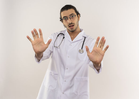 停止戴着光学眼镜的年轻男医生穿着白色长袍 听诊器在隔离的白色墙壁上显示停止手势 并留有复印空间长袍男性眼镜