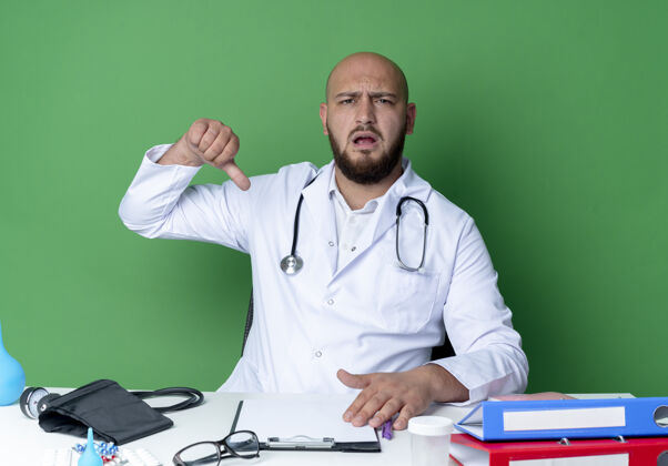 听诊器年轻的男医生穿着医用长袍 手持听诊器坐在办公桌旁 手里拿着医疗工具 他的大拇指被隔离在绿色的墙上拇指医疗年轻