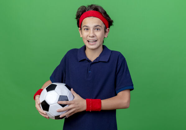男孩令人印象深刻的年轻英俊的运动男孩戴着头带和戴着牙套的护腕拿着足球看着相机隔离在绿色背景与复制空间头带腕带运动