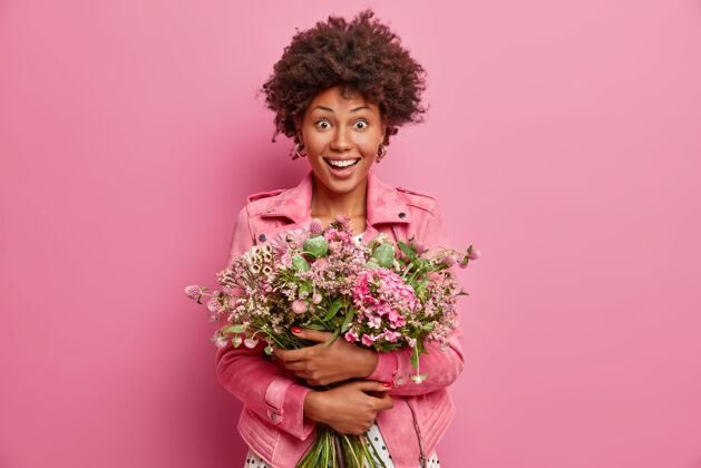 特别喜出望外的黑皮肤女人抱着一束漂亮的花 去跟朋友祝贺结婚纪念日 穿着粉色的时尚夹克 站在室内庆祝 特别的场合春天夹克人