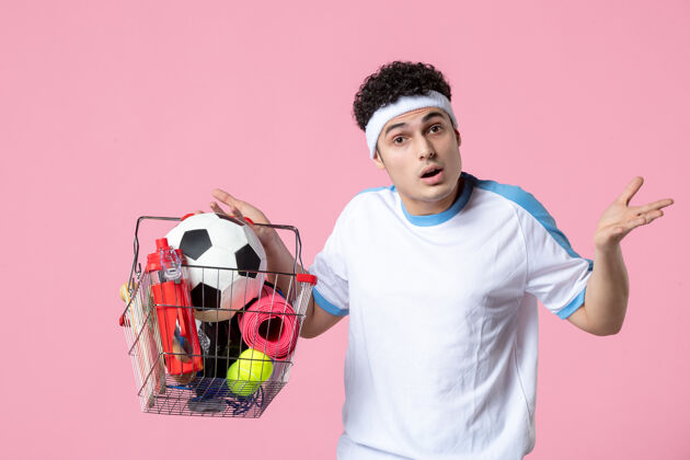 前面正面图穿着运动服的年轻男子 带着装满运动用品的篮子 粉色的墙壁人年轻男性健身房