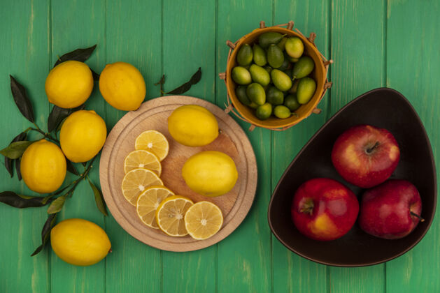 切片木制厨房板上的柠檬片俯视图 碗上放着红苹果 桶上放着金盏花 柠檬被隔离在绿色的木墙上板木头柑橘