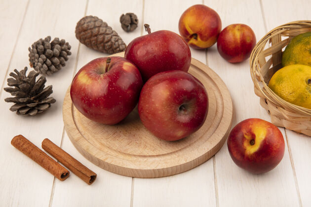 桶顶视图：新鲜苹果放在木制厨房板上 橘子放在桶上 桃子 肉桂 松果孤立在白色的木墙上顶部橘子等级