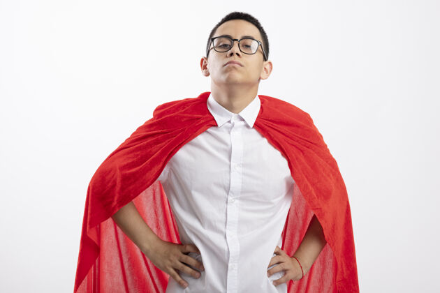 男孩穿着红斗篷的自信的年轻超级英雄男孩戴着眼镜看着相机 手放在白色背景下的腰部手穿白色