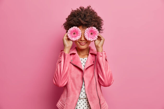 乐趣年轻的卷发女子手持粉色非洲菊雏菊 遮住眼睛 穿着时尚的粉色夹克 做装饰 在室内摆姿势乐观花举行