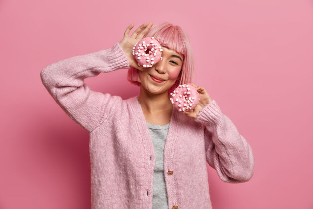 心情积极微笑的亚洲女人有乐趣 拿着两个美味的甜甜圈 玩甜品 享受开胃甜点 穿着休闲套头衫 甜点发型甜甜圈
